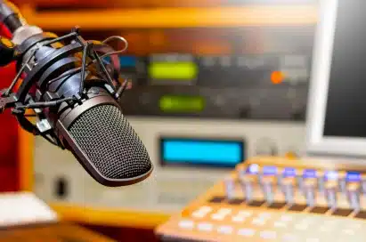 Close-up of a professional microphone in a recording studio, with audio mixing equipment and blurred control panel in the background.
