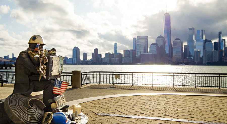 A bronze firefighter statue with memorial items faces the New York City skyline across the river on a partly cloudy day.