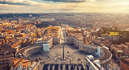 Aerial view of St. Peter's Square in Vatican City, featuring the central obelisk, colonnades, and surrounding cityscape under a partly cloudy sky.