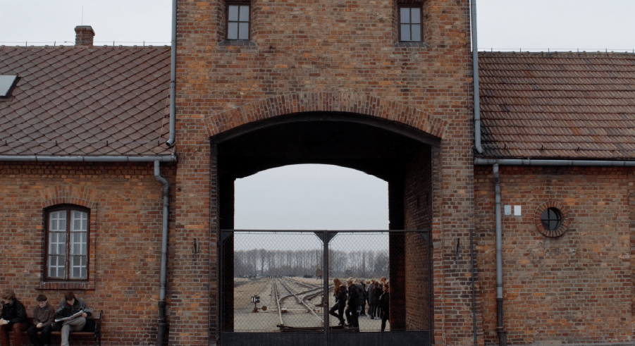 Brick entrance gate of Auschwitz concentration camp with people standing inside and outside, railway tracks visible through the archway.