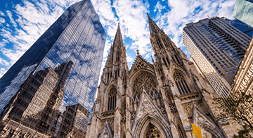 Gothic-style cathedral with two spires stands between modern glass and concrete skyscrapers under a partly cloudy sky.