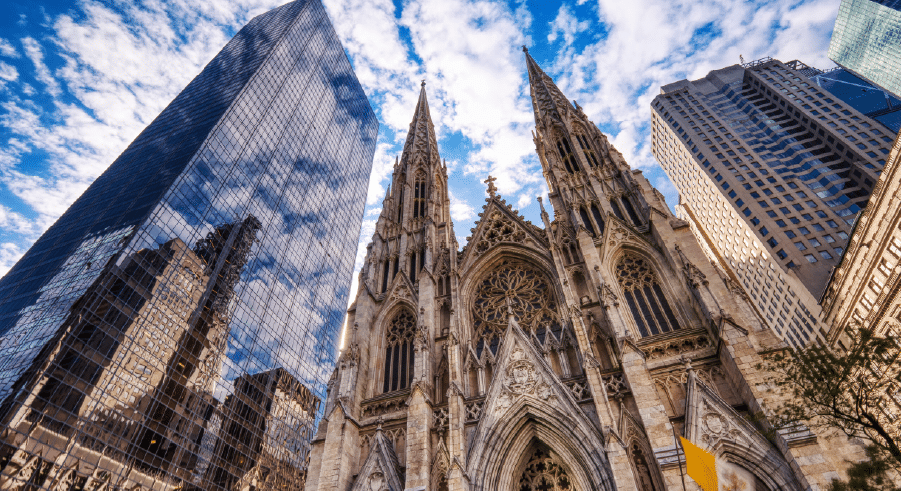 Gothic-style cathedral with pointed arches and spires stands between modern glass and concrete skyscrapers under a partly cloudy sky.