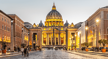 View of St. Peter's Basilica in Vatican City at dusk, with people walking along the cobbled street and the building illuminated by lights.