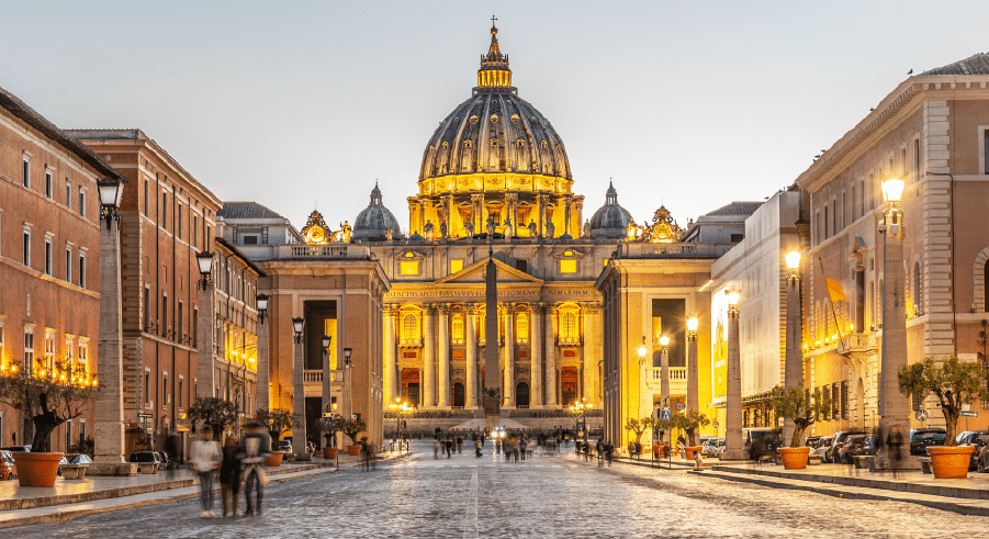 St. Peter's Basilica in Vatican City is illuminated at dusk, with people walking along the cobbled street leading to the entrance.