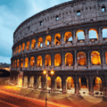 The Colosseum in Rome is illuminated at dusk, with blurred car lights passing by on the street in the foreground.