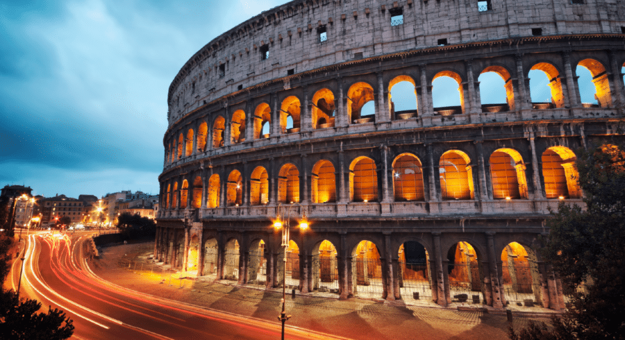 The Colosseum in Rome is illuminated at dusk, with light trails from passing vehicles visible on the street beside the ancient amphitheatre.