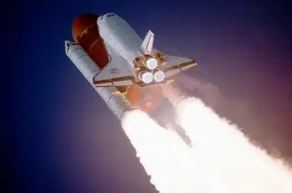 A space shuttle launches into the sky with bright flames and smoke trailing from its rocket boosters against a dark blue background.