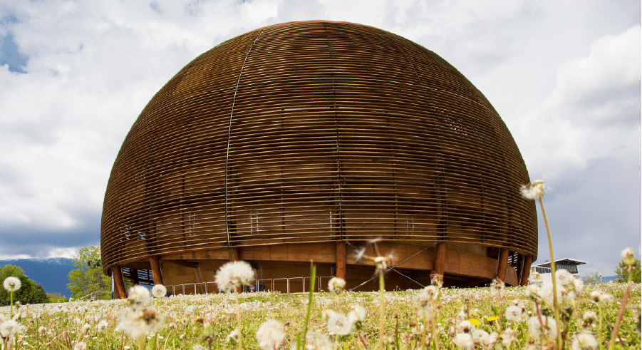 A large, spherical wooden building stands in a grassy field with dandelions under a cloudy sky—an intriguing spot for science trips in Iceland.