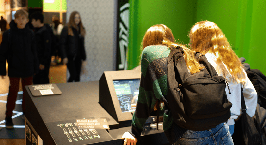 Two people with long hair and rucksacks interact with a touchscreen display in a brightly lit indoor space with green walls, capturing the spirit of science trips London is known for. Other people walk in the background.