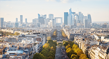 A wide view of a cityscape featuring a tree-lined avenue leading towards modern skyscrapers and the Grande Arche in the distance.
