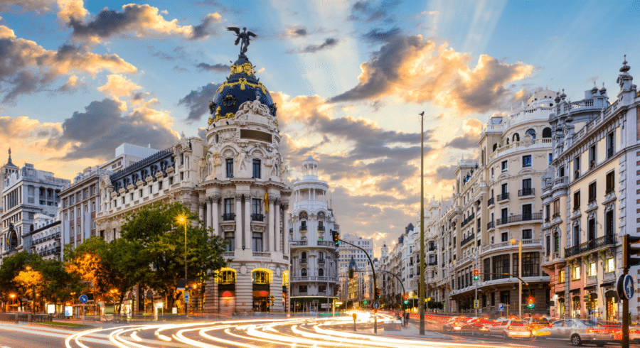 A busy street scene in Madrid featuring the historic Metropolis Building with ornate architecture, illuminated by city lights at sundown.