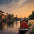 A narrowboat is moored along a canal at sunset, with buildings on the left and trees and grass on the right.