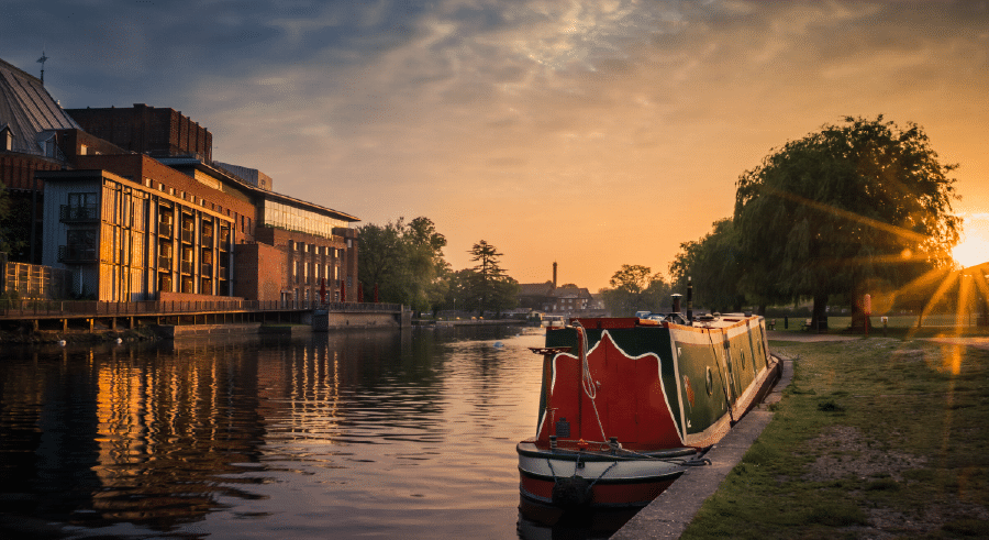 A canal boat is moored on a calm river at sunrise, with buildings and trees reflected in the water and the sun low on the horizon.
