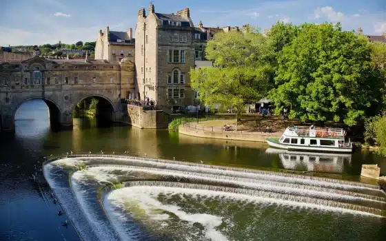 Stone bridge and historic buildings beside a river with a weir; a tour boat is moored near trees on a sunny day.