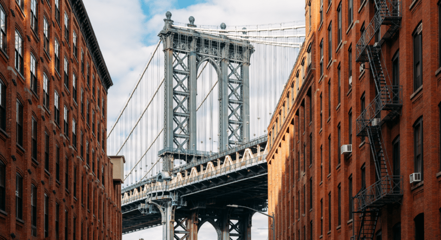 A view of the Manhattan Bridge framed by two red brick buildings, with a portion of the Empire State Building visible through the bridge structure.