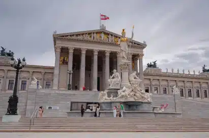 The Austrian Parliament Building with statues and steps in front, the Austrian flag flying on the roof, and several people visible near the entrance.