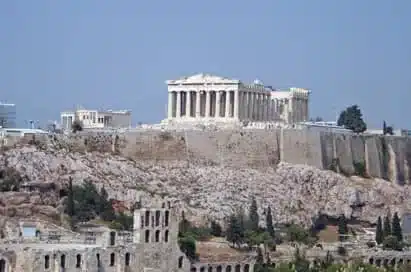 The Parthenon stands atop the Acropolis in Athens, Greece, with ancient ruins and rocky terrain in the foreground under a clear sky.