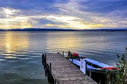 A wooden jetty extends into a calm lake under a cloudy sky at sunset, with several small boats moored alongside the jetty.