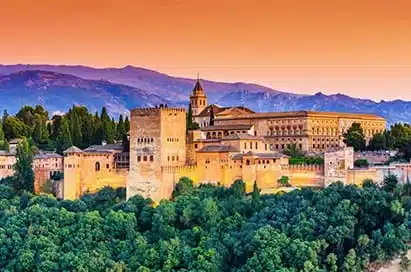 The Alhambra palace complex in Granada, Spain, is shown at sunset with mountains in the background and trees in the foreground.