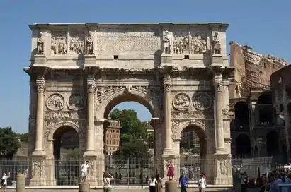The Arch of Constantine, a large Roman triumphal arch with three archways, stands in front of the Colosseum in Rome, with tourists nearby on a sunny day.