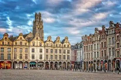 Historic European square with ornate, gabled buildings and a tall church tower under a partly cloudy sky; cobbled plaza in the foreground.