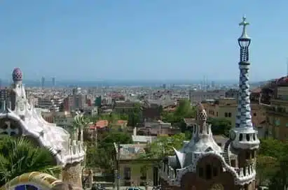 View of Barcelona cityscape from Park Güell, featuring distinct mosaic architecture in the foreground and the Mediterranean Sea in the distance under a clear sky.