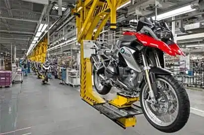 A motorcycle is suspended on a yellow assembly line inside a large, organised factory with additional motorcycles and equipment visible in the background.