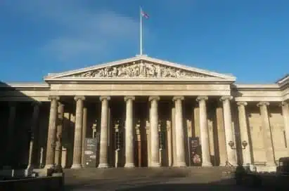 The façade of a neoclassical building with tall columns, a triangular pediment, and a flag on top under a clear blue sky.