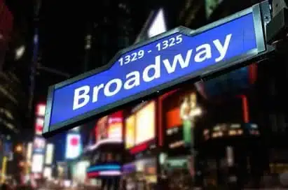 Blue street sign for "Broadway" with building numbers 1329–1325, illuminated at night with blurred city lights and hoardings in the background.