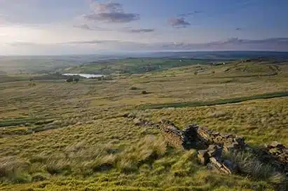 Rolling green hills with patches of grass, a stone wall in the foreground, and a distant body of water under a cloudy sky.