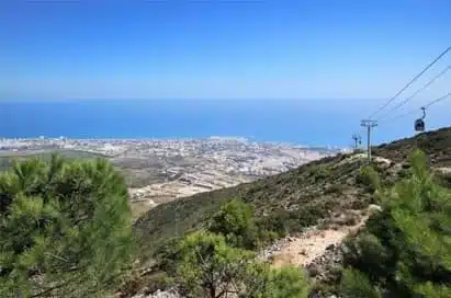 View from a hillside with green shrubs, overlooking a coastal city and the sea; a cable car system runs up the hill on the right side under a clear blue sky.