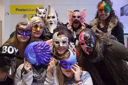 A group of people pose indoors wearing colourful and uniquely designed masks, obscuring their faces. A yellow "Poste Italiane" sign is visible in the background.