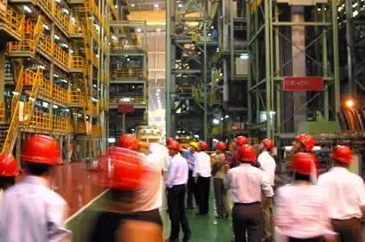 A group of people wearing red hard hats tour a large industrial facility with metal structures and yellow railings.