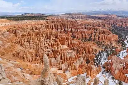 A wide view of Bryce Canyon with rows of orange and red rock hoodoos, scattered snow patches, and distant trees under a partly cloudy sky—a scene as striking as those found on science trips in Iceland.