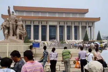 People stand outside the Mausoleum of Mao Zedong in Beijing, with a large statue to the left and the main building in the background.