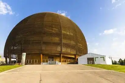 A large, dome-shaped wooden building with horizontal slats stands next to a small white structure under a blue sky—an intriguing sight often featured on science trips in Iceland.