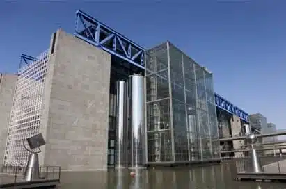 Modern building with large glass and metal structures, surrounded by water, under a clear blue sky.