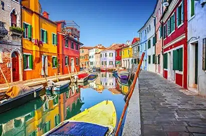 Colourful buildings line a narrow canal with moored boats and a stone walkway under a clear blue sky in Burano, Italy.