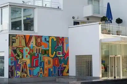 Colourful mural with overlapping, stylised letters on the side of a modern white building with large windows and balconies.