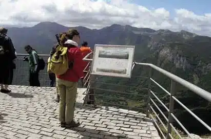 Several people stand on a mountain viewing platform with a railing, looking at the scenery and an information sign. Distant mountains and cloudy sky are visible.