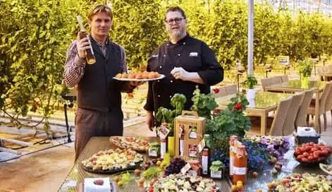 Two men stand by a table filled with assorted food and drinks in an indoor glasshouse setting with rows of plants in the background.