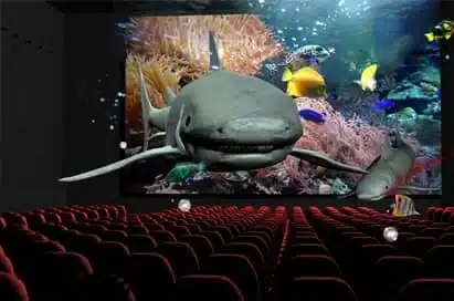 A nearly empty cinema showing a 3D underwater scene with a shark and fish appearing to come out of the screen towards the seats.
