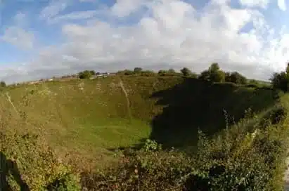 A large grassy crater with sloping sides, surrounded by vegetation and trees under a partly cloudy sky.