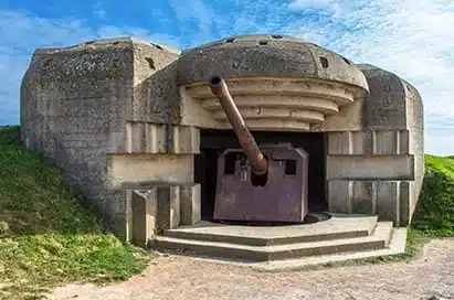 A concrete Second World War bunker with a large artillery gun positioned in the centre, set against a grassy landscape under a partly cloudy sky.