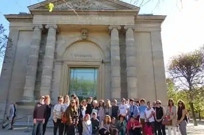 A group of people stands in front of a historic stone building with columns and an arched entrance on a sunny day.