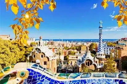 View of Park Güell in Barcelona, featuring colourful mosaic tiles in the foreground, whimsical architecture, and a cityscape with the sea in the background on a sunny day.