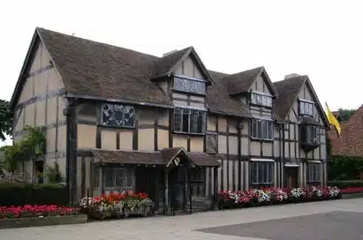 A large, historic timber-framed house with leaded glass windows, flowerbeds in front, and a yellow flag on the right-hand side.