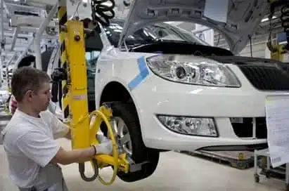A factory worker operates machinery to fit a wheel to a white car on an automotive assembly line.