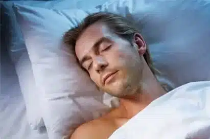 A man with fair hair is lying in bed, asleep on a white pillow and covered with a white sheet.