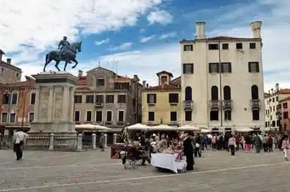 A public square with market stalls, people gathered, historic multi-storey buildings, and an equestrian statue under a partly cloudy sky.